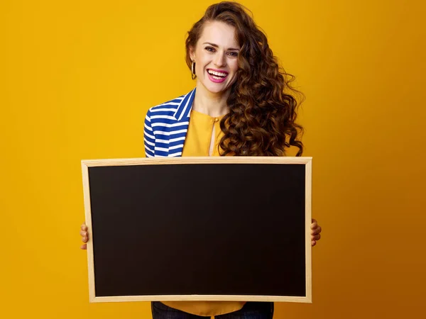 空白ボードを示す黄色の背景に長い波状のブルネットの髪を持つスタイリッシュな女性を笑顔 — ストック写真