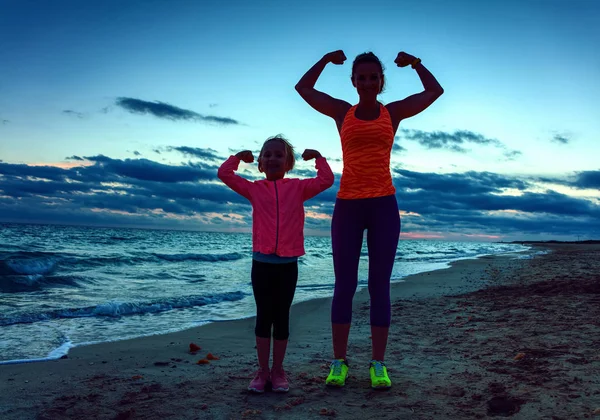 healthy mother and child in sport style clothes on the seacoast on the sunset showing biceps