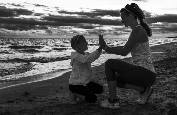 young mother in sport style clothes giving child high five on the seacoast on the sunset