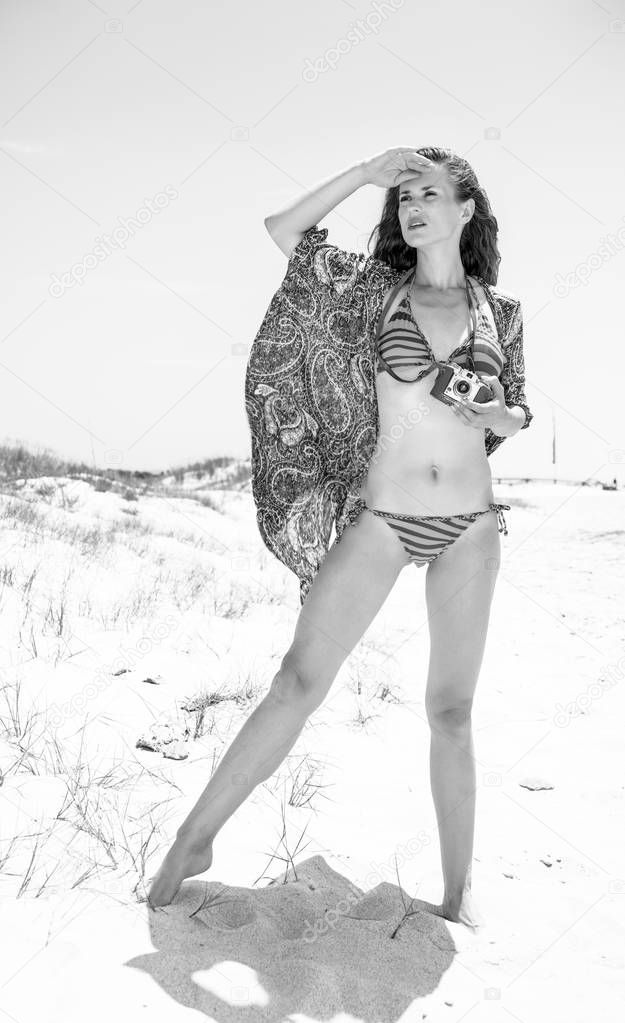 Full length portrait of bohemian young woman in swimwear on the seacoast with retro film camera looking into the distance