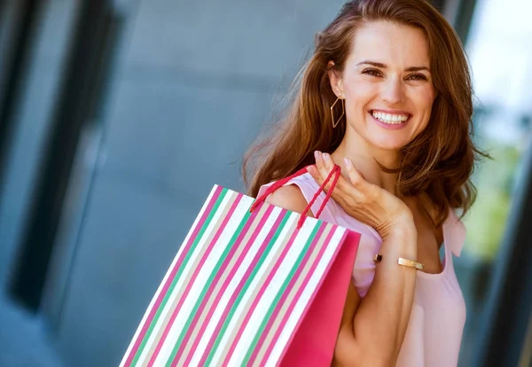 A brown-haired woman is seen from the waist up. She holds a striped, multi-coloured and red shopping bag over her right shoulder. Her smile is relaxed and happy. She is effortlessly stylish and classic. Her wide smile make her approachable, happy, an