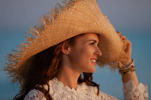 Sonriente mujer en la orilla del mar al atardecer mirando a la distancia — Foto de Stock