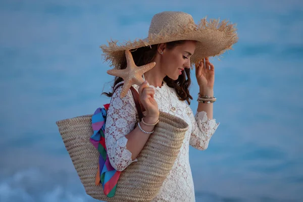 Mujer en la playa al atardecer sosteniendo playa bolsa de paja y estrellas de mar — Foto de Stock