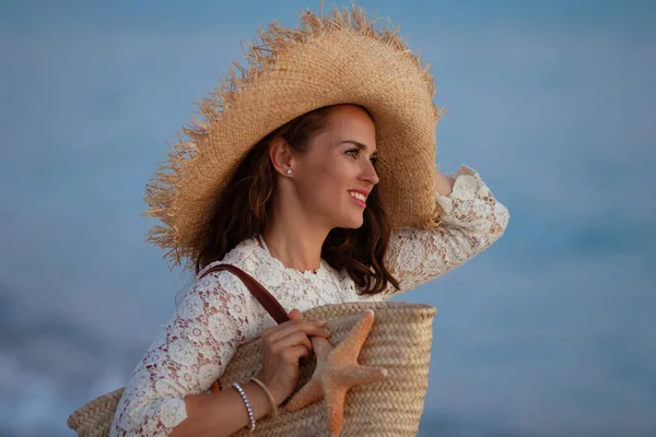 Femme sur la plage au coucher du soleil tenant sac de paille de plage et étoile de mer — Photo