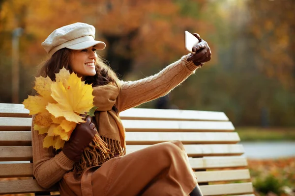 Sorrindo mulher moderna com folhas amarelas tirando foto com telefone — Fotografia de Stock