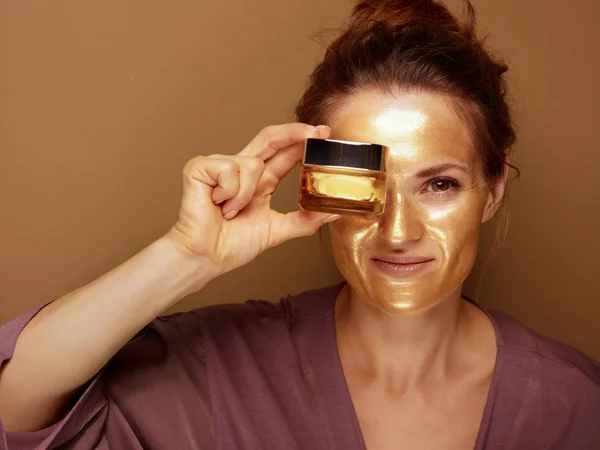 Kobieta ze złotą maską pokazuje krem na brązowym tle — Zdjęcie stockowe