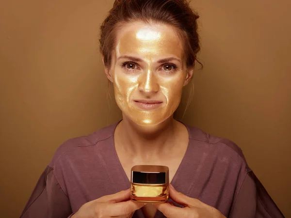 Elegancka kobieta ze złotą maską trzymająca butelkę kremu do twarzy — Zdjęcie stockowe