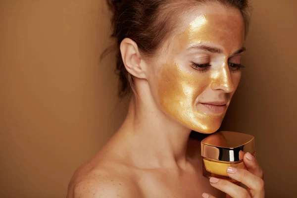 Женщина в золотой маске держит крем на коричневом фоне — стоковое фото