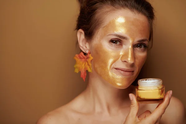 Huisvrouw met gouden masker houden crème op bruine achtergrond — Stockfoto