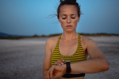 Gün batımında deniz kıyısında spor kıyafetleri içindeki formda bir kadının portresi. Adımları takip etmek, koşmak veya spor uygulamasında sürmek için akıllı saati kullanıyor..