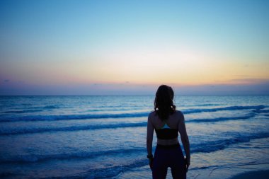 Seen from behind fit woman in sport clothes on the seacoast in the evening relaxing after workout.