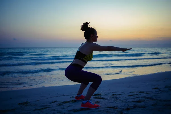 フィットネススポーツ女性のシルエットフィットネス服でビーチで夜にスクワットをやって — ストック写真