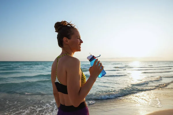 young woman in fitness clothes with bottle of water on the ocean coast at sunset.