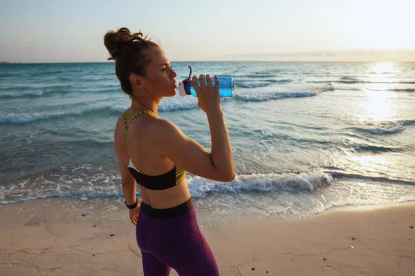fitness woman in fitness clothes on the beach in the evening drinking water from bottle.