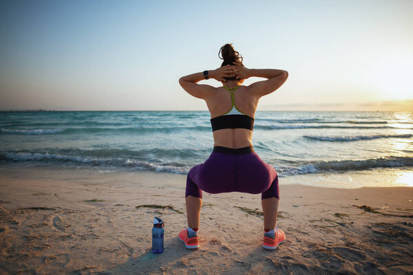 Seen from behind active woman in sport style clothes on the seashore in the evening doing squats.