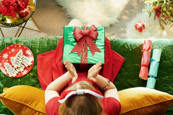 Holiday Season. Upper view of smiling modern little princess in red dress with Christmas present box clapping while sitting on couch in the modern living room at Christmas.