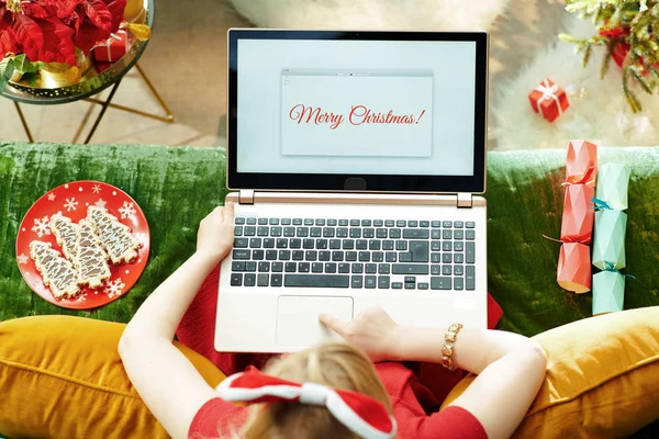 假期季节 圣诞前夕 身穿红色礼服的现代小公主坐在现代客厅的长椅上 用笔记本电脑写着圣诞快乐的电子邮件 — 图库照片