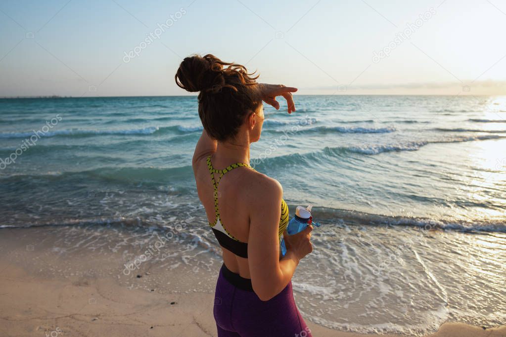 Seen from behind healthy sports woman in fitness clothes with bottle of water looking into the distance on the seashore in the evening.