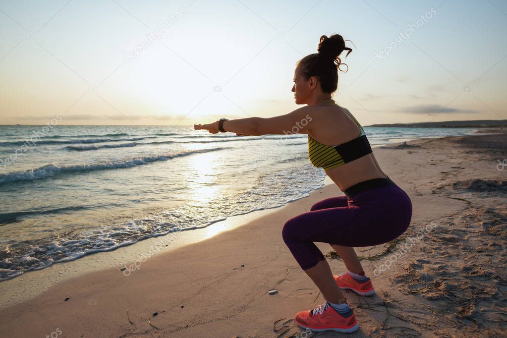 healthy woman in sport clothes on the seashore in the evening doing squats.