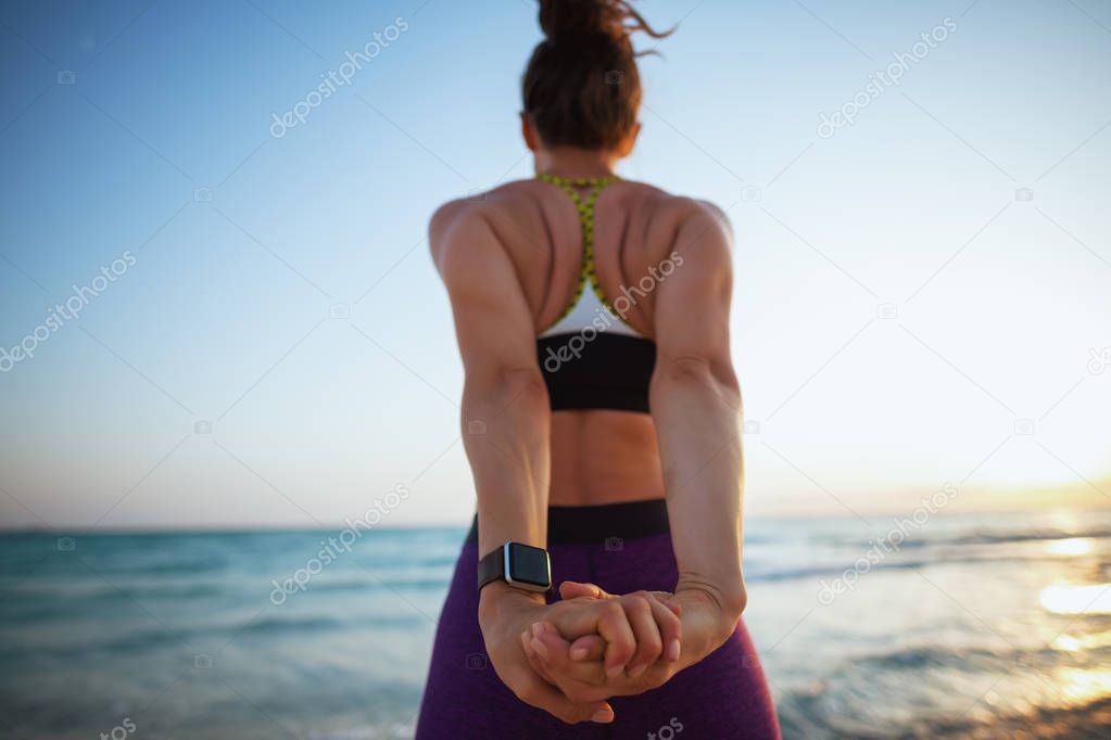 Seen from behind young sports woman in fitness clothes on the beach at sunset stretching.