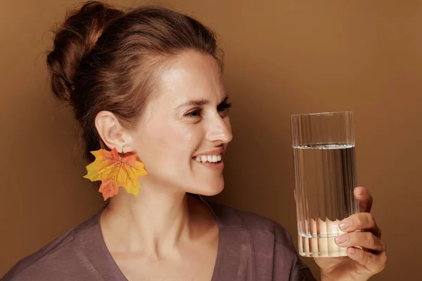 こんにちは秋 茶色の背景に水のガラスを見て秋の葉のイヤリングとバスローブで笑顔の中年女性の肖像画 — ストック写真