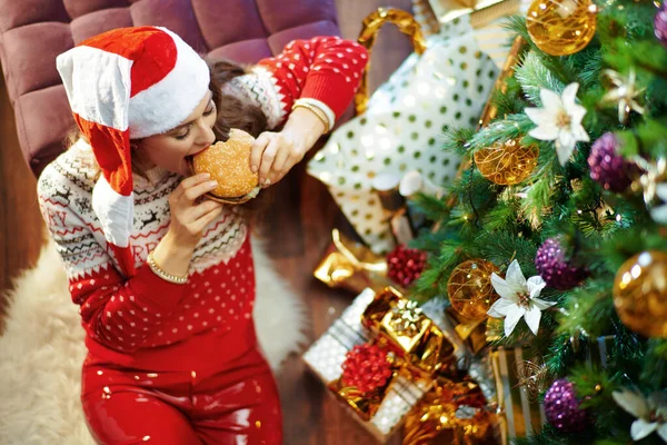 上のビューの笑顔現代40歳の女性とともに長いブルネットの髪を身に着けている赤白クリスマスセーターと自己サンタ帽子下装飾クリスマスツリー近く現在のボックスかむハンバーガー — ストック写真