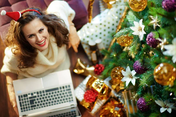 上のビューの幸せなトレンディーな中年女性とともに長いブルネットの髪でゴールドスパンコールスカートとホワイトセーター下装飾クリスマスツリー近く現在のボックスを使用してラップトップ — ストック写真