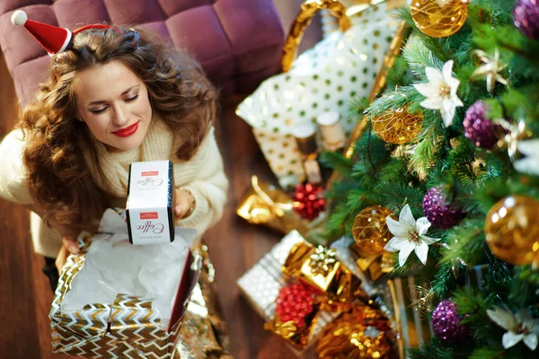 在装饰过的圣诞树下 穿着金色亮片裙 头戴白色毛衣 留着一头长长的黑发的家庭主妇微笑着 她站在装有撕破的圣诞礼盒和咖啡香味的礼盒旁边 — 图库照片