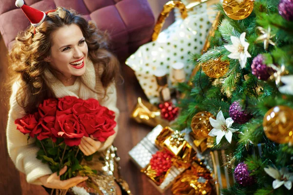 在装饰过的圣诞树下的礼物盒旁边 穿着金黄色亮片裙和红色玫瑰白色毛衣的时髦家庭主妇微笑着 看着装饰过的圣诞树下的复制空间 — 图库照片