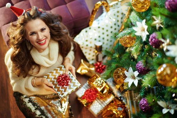 上のビューの笑顔スタイリッシュな女性とともに長いブルネットの髪でゴールドスパンコールスカートと白セーター下装飾クリスマスツリー近く現在のボックス保持ラップ本 — ストック写真