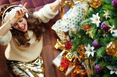 Uzun kahverengi saçlı, altın rengi etekli, beyaz kazaklı, kulaklıklı ve hediye kutularının yanında süslü Noel ağacının altında dans eden mutlu ve modaya uygun bir kadının üst görüntüsü..