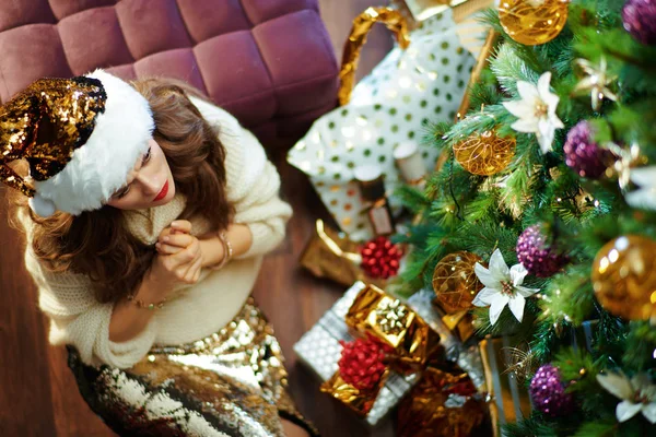 忧心忡忡的中年家庭主妇 长长的黑发 金色亮片裙 白色毛衣 挂在装饰过的圣诞树下 靠近地板上的盒子 — 图库照片