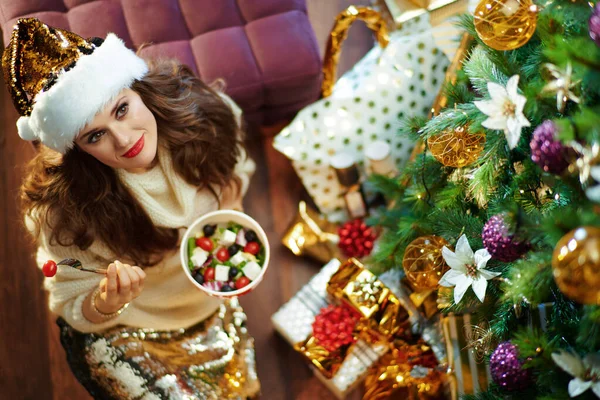 在装饰圣诞树下的金色亮片裙和白色毛衣中 现代女人有着一头长长的黑发 正在吃着健康的沙拉 — 图库照片