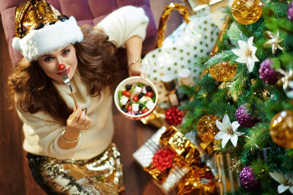 上图是快乐的现代女性 一头长长的黑发 穿着金色亮片裙 白色毛衣 配上健康的沙拉 在装饰过的圣诞树下用樱桃番茄做红鼻子 放在礼品盒旁 — 图库照片