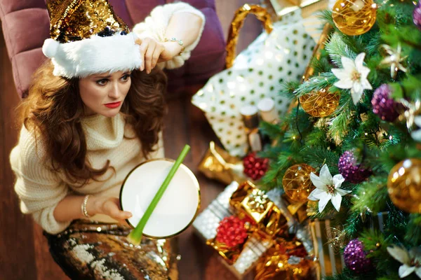 身穿金黄色亮片裙 头戴白色毛衣 留着芹菜盘 装饰过的圣诞树下靠近礼物盒的不高兴的年轻女子的上图 — 图库照片