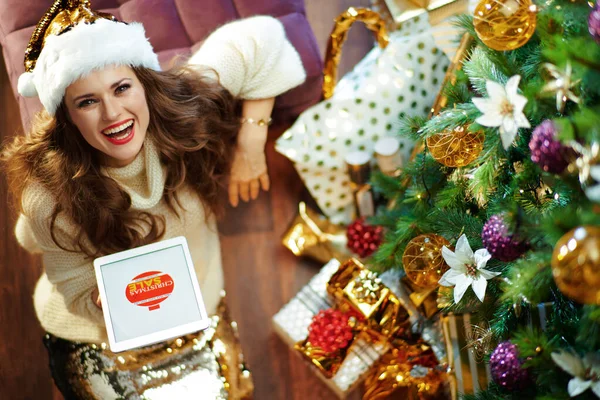在礼物盒附近装饰过的圣诞树下 身穿金色亮片裙 头戴白色毛衣 面带微笑的女人的上图在平板电脑Pc上得到了优惠券 — 图库照片