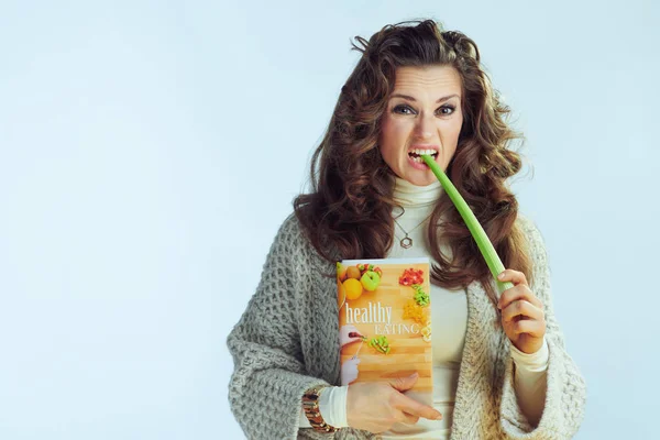 Unhappy young housewife with healthy eating book eating celery — 스톡 사진