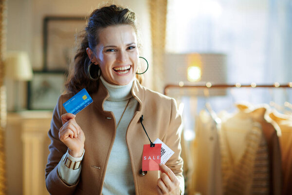 smiling trendy middle age woman in white sweater and skirt trying beige coat with red sale price tag and holding blue credit card in modern fashion store.
