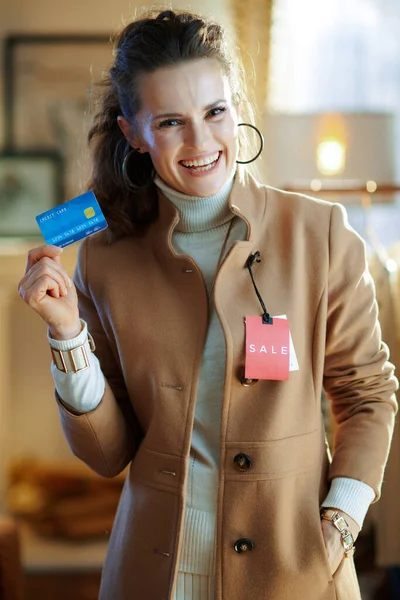 白いセーターとスカートの若い女性の笑顔試すベージュコートとともに赤販売価格タグと保持青クレジットカードで現代ファッション店 — ストック写真