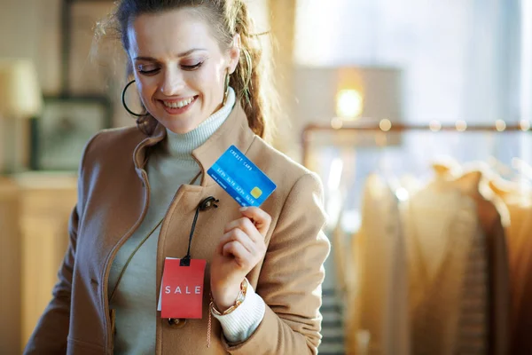 白いセーターとスカートの若い女性の笑顔試すベージュコートとともに赤販売価格タグと保持青クレジットカードで現代ファッションブティック — ストック写真