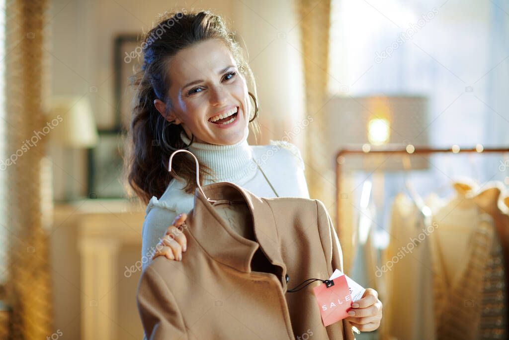 happy elegant woman in white sweater and skirt showing beige coat with sale price tag in modern fashion store.