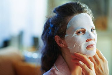 Güneşli kış gününde modern evde 40 yaşında pijamalı bir kadın beyaz bir maske takıyor..