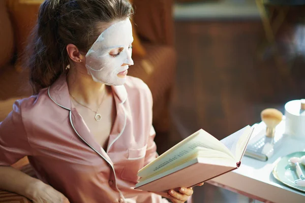 スタイリッシュな40歳の主婦ですパジャマにホワイトシートフェイスマスク読書本で現代の家で晴れた冬の日 — ストック写真