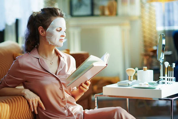 若いです女性でパジャマでホワイトシートフェイスマスク読書本で現代の家で晴れた冬の日 — ストック写真