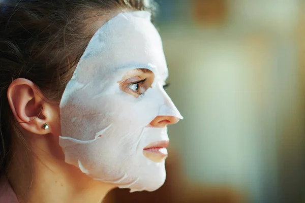 Side view of modern 40 years old woman in pajamas with white sheet facial mask on face in the modern living room in sunny winter day.