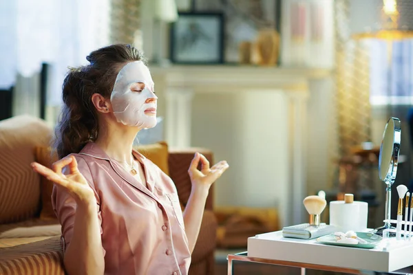 リラックスしたエレガントな女性でパジャマとともにホワイトシート顔マスク上の顔瞑想で現代の家で晴れた冬の日 — ストック写真