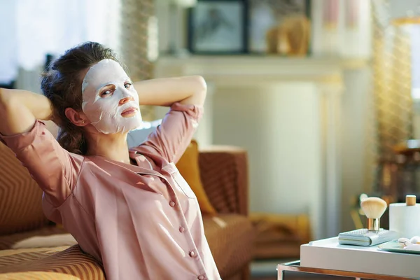 白いシート顔のマスクを持つパジャマでリラックスしたエレガントな中年女性の肖像画は 晴れた冬の日に現代的な家の中で顔に — ストック写真