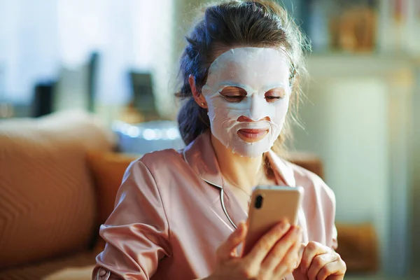 エレガントな中年女性でパジャマとともにホワイトシートフェイシャルマスク上の顔見ビデオでインターネット経由スマートフォンで現代のリビングルームで晴れた冬の日 — ストック写真