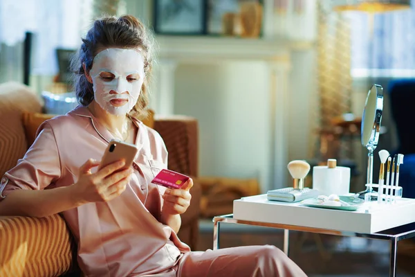 modern woman in pajamas with white sheet facial mask on face holding smartphone with credit card browsing online shop in the modern living room in sunny winter day.