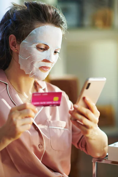 在阳光明媚的冬日里 身穿睡衣 头戴白色口罩 手持智能手机 头戴信用卡的40岁时髦女性在现代客厅里上网购物 — 图库照片